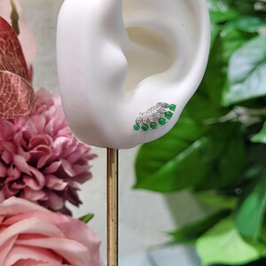 May include: A silver and green earring is displayed on a white ear-shaped form. The earring features a fan-shaped design with small, round, green gemstones and clear accent stones. The earring is set against a backdrop of pink flowers and green foliage.