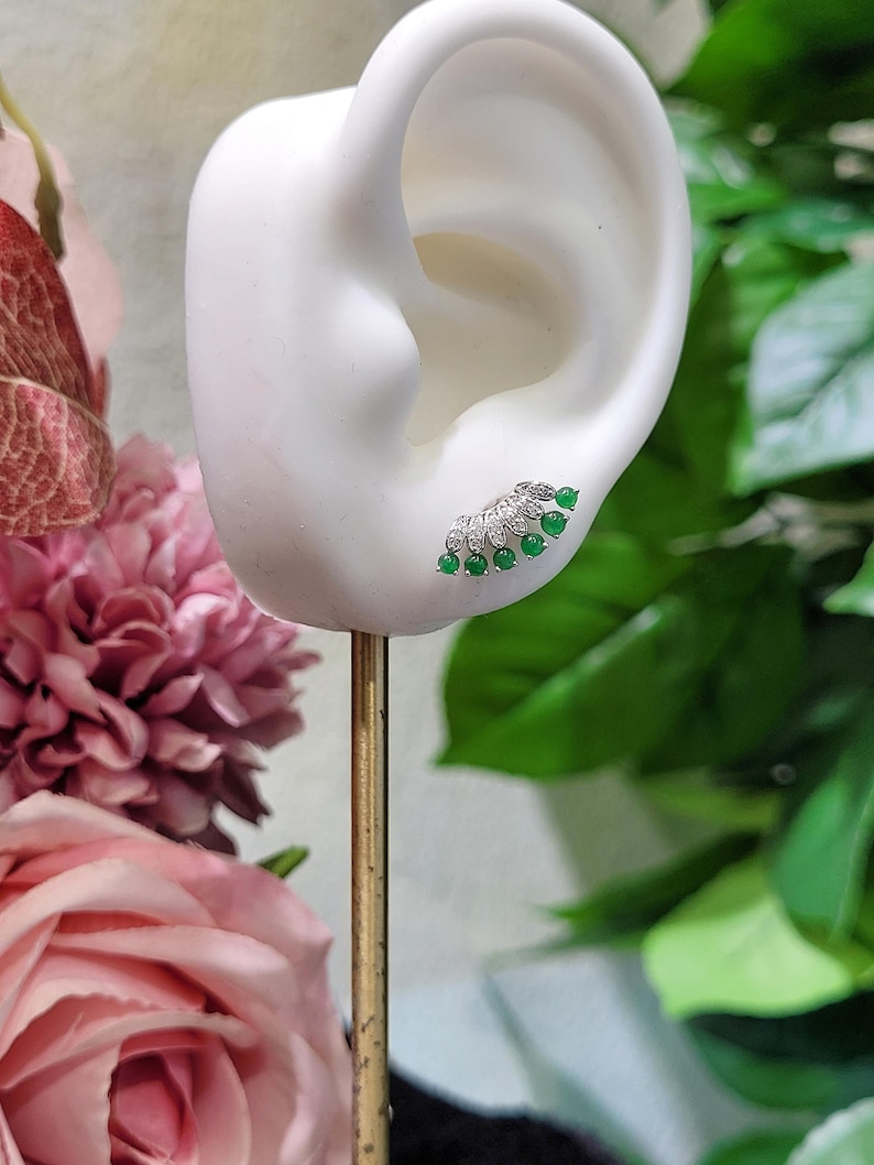 May include: A silver and green earring is displayed on a white ear-shaped form. The earring features a fan-shaped design with small, round, green gemstones and clear accent stones. The earring is set against a backdrop of pink flowers and green foliage.