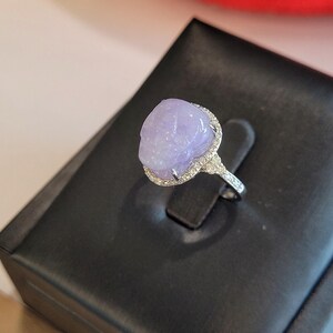 May include: A silver ring featuring a large, oval-shaped, lavender-colored gemstone. The gemstone is surrounded by a halo of small, clear stones. The ring is displayed on a black, textured surface.