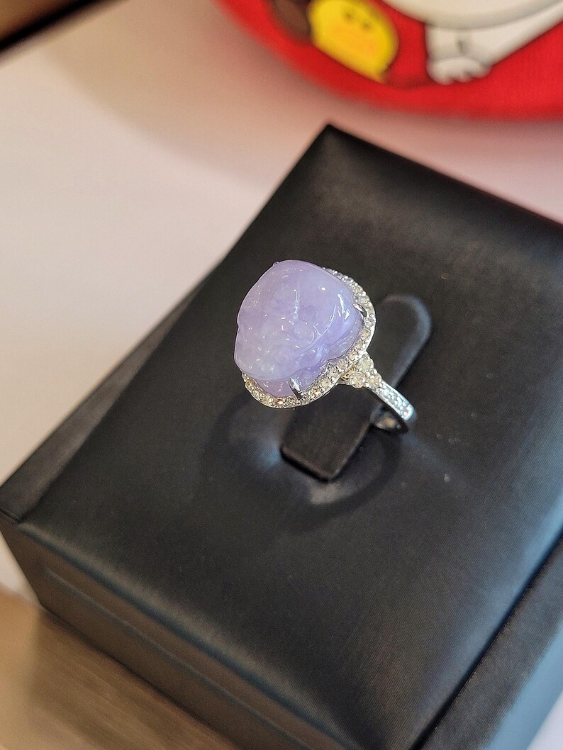 May include: A silver ring featuring a large, oval-shaped, lavender-colored gemstone. The gemstone is surrounded by a halo of small, clear stones. The ring is displayed on a black, textured surface.