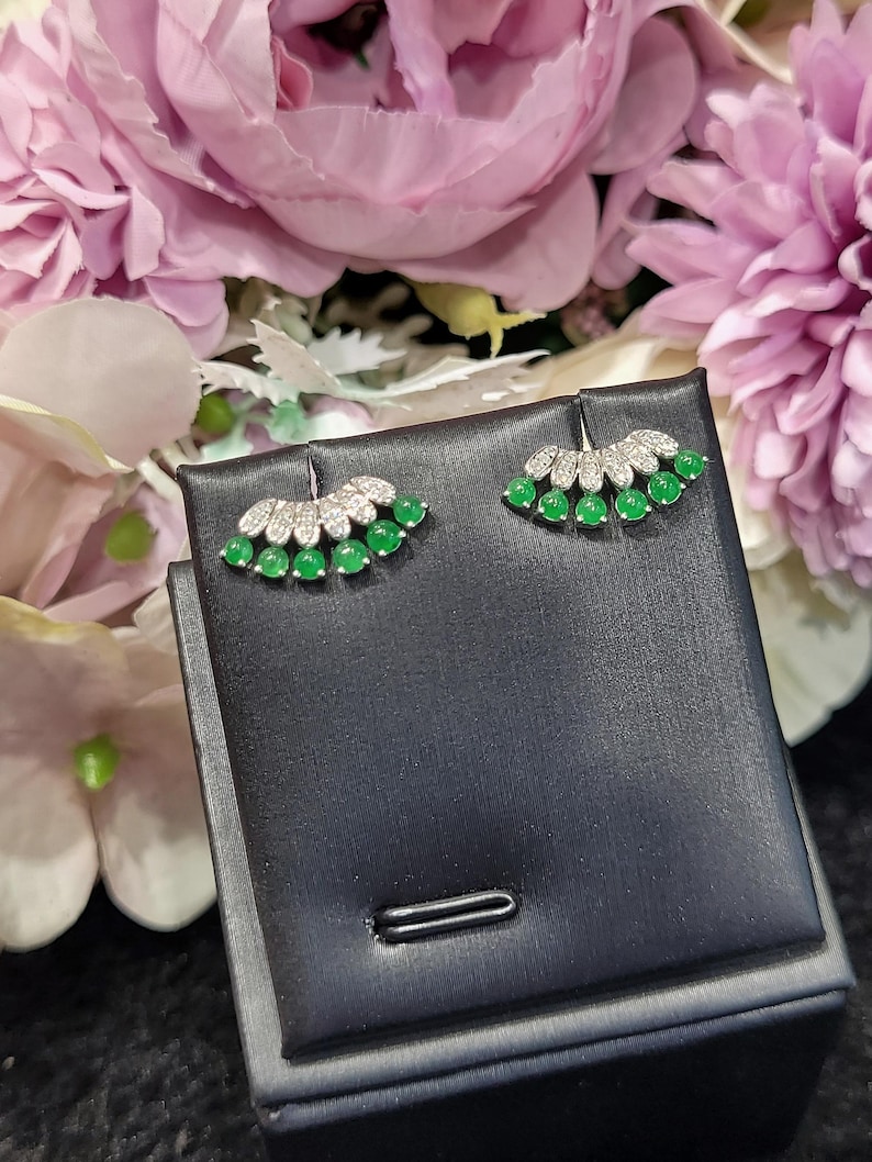 May include: A pair of silver earrings with a fan-shaped design. Each earring features a row of small, round, bright green gemstones, topped with a section of clear, sparkling stones. The earrings are displayed on a black jewelry box, with a backdrop of pink and purple flowers.