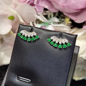 May include: A pair of stud earrings featuring a fan-shaped design. Each earring has a row of small, round, bright green gemstones and a top row of clear, sparkling stones. The earrings are displayed on a black surface against a backdrop of pink and white flowers.