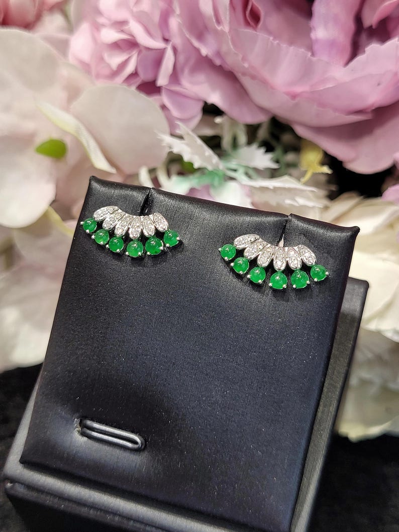 May include: A pair of stud earrings featuring a fan-shaped design. Each earring has a row of small, round, bright green gemstones and a top row of clear, sparkling stones. The earrings are displayed on a black surface against a backdrop of pink and white flowers.