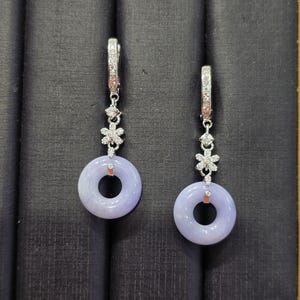 May include: A pair of silver earrings with a floral design and lavender-colored circular jade pendants. The earrings have a post-and-clip closure and are displayed against a dark background. The jade pendants are approximately 1 inch in diameter.