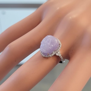May include: A close-up of a ring featuring a large, oval-shaped, lavender-colored gemstone. The gemstone is surrounded by a halo of small, clear gemstones set in a silver band. The ring is on a person's finger.