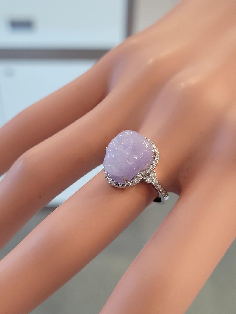 May include: A close-up of a ring featuring a large, oval-shaped, lavender-colored gemstone. The gemstone is surrounded by a halo of small, clear gemstones set in a silver band. The ring is on a person's finger.