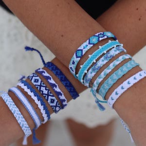 May include: A collection of handmade friendship bracelets in shades of blue and white. The bracelets feature various woven patterns, including diamonds, stripes, and hearts. The bracelets are worn on crossed arms, showcasing the different designs and colors.
