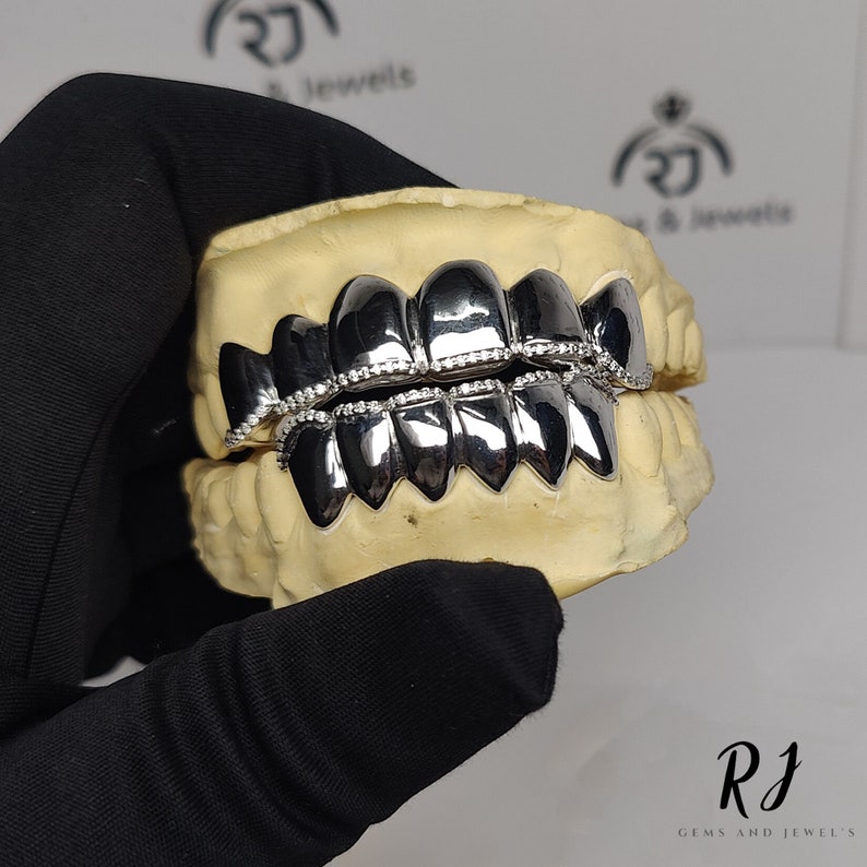 May include: A silver grill with a row of small diamonds around the edge. The grill is displayed on a model of a mouth.