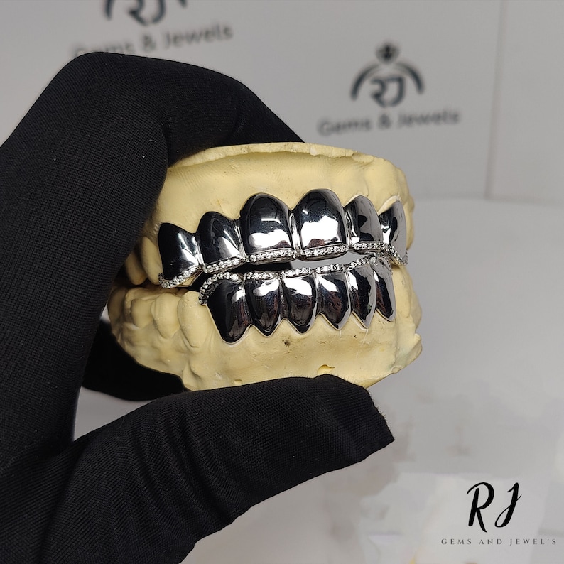 May include: A silver grill with a row of small diamonds around the edge. The grill is displayed in a dental mold.