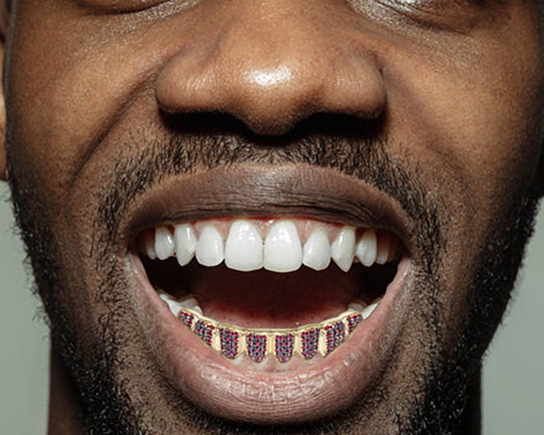 May include: A close-up of a person's mouth with a gold grill with red jewels. The person is smiling and showing their teeth.