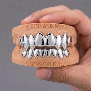 Puede incluir: Un conjunto de fundas dentales de metal color plata ajustadas sobre un modelo dental de color beige. El texto RJ GEMS AND JEWELS está grabado en las secciones de las encías superior e inferior del modelo. Las fundas tienen un acabado liso.