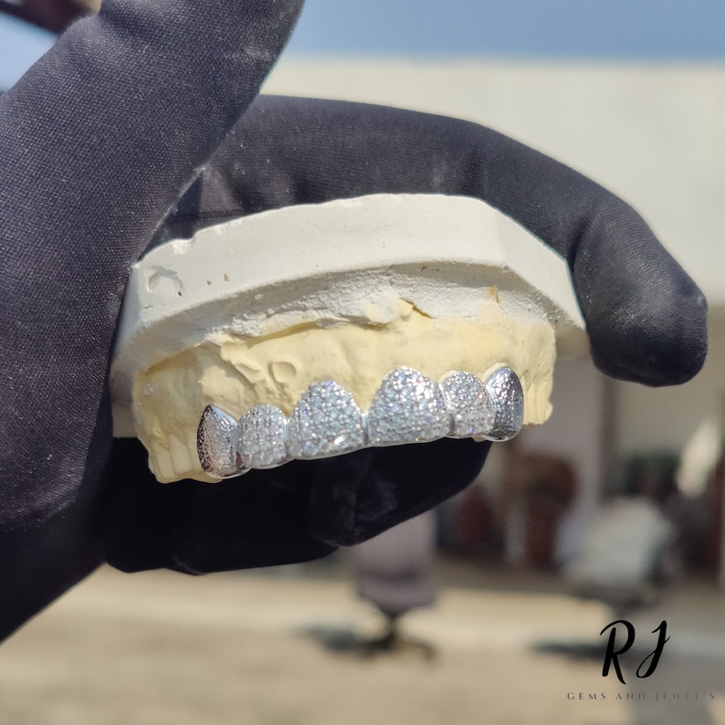 May include: A set of silver grillz with a row of small, clear stones. The grillz are set in a mold of a mouth.