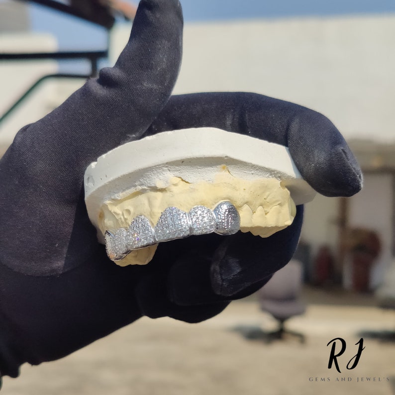 May include: Silver grill with a row of clear cubic zirconia stones. The grill is displayed on a white mold of a mouth.