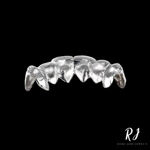 May include: Silver grill with five teeth, designed to fit over the front teeth. The grill is made of a smooth, shiny metal.