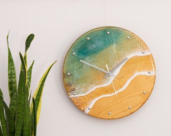 May include: A round wooden clock with a resin ocean scene. The clock face features a blue-green ocean with white waves crashing on a sandy beach. The clock hands are silver.