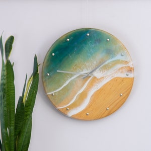 Peut inclure: Une horloge ronde en bois avec une scène océanique en résine. Le cadran de l'horloge présente un océan bleu et vert avec des vagues blanches qui se brisent sur une plage de sable. Les aiguilles de l'horloge sont argentées.