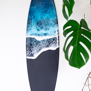 May include: A black surfboard with a blue and white resin ocean wave design. The surfboard is hanging on a white wall.