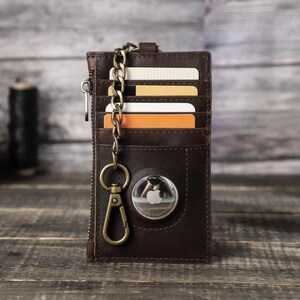 Puede incluir: Una cartera de cuero marr&oacute;n con cierre de cremallera y una cadena de metal. La cartera tiene m&uacute;ltiples ranuras para tarjetas y un peque&ntilde;o bolsillo para efectivo. La cartera tambi&eacute;n tiene un c&iacute;rculo de metal con un logotipo de Apple.