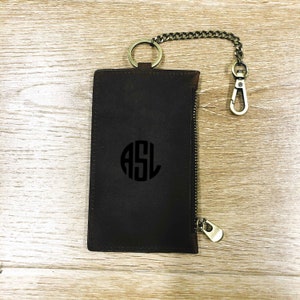 Puede incluir: Una cartera de cuero marr&oacute;n con cremallera, una cadena de metal y un llavero. La cartera tiene un c&iacute;rculo con monograma con las iniciales "ASL" en negro.