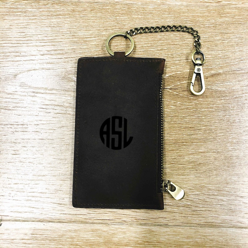 Puede incluir: Una cartera de cuero marr&oacute;n con cremallera, una cadena de metal y un llavero. La cartera tiene un c&iacute;rculo con monograma con las iniciales "ASL" en negro.