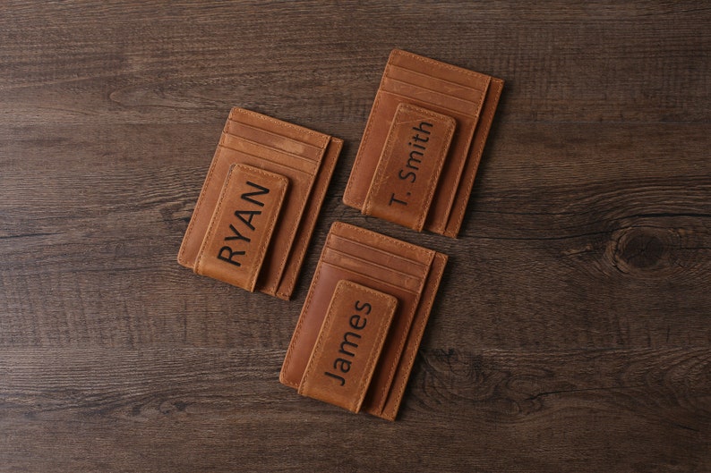 May include: Three brown leather wallets with a money clip. The wallets have the names "RYAN", "James", and "T. Smith" engraved on them.