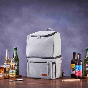 May include: A gray insulated backpack cooler with two zippered compartments and a brown leather patch. The cooler is on a wooden table with various bottles of beer and a glass of ice.