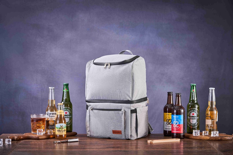May include: A gray insulated backpack cooler with two zippered compartments and a brown leather patch. The cooler is on a wooden table with various bottles of beer and a glass of ice.