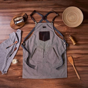 K&ouml;nnte beinhalten: Eine graue Leinensch&uuml;rze mit braunen Ledereins&auml;tzen und verstellbaren Tr&auml;gern. Die Sch&uuml;rze hat eine gro&szlig;e Tasche auf der Vorderseite und zwei kleinere Taschen an den Seiten.