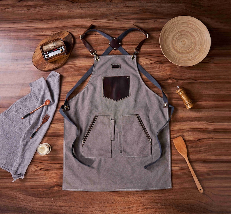 K&ouml;nnte beinhalten: Eine graue Leinensch&uuml;rze mit braunen Ledereins&auml;tzen und verstellbaren Tr&auml;gern. Die Sch&uuml;rze hat eine gro&szlig;e Tasche auf der Vorderseite und zwei kleinere Taschen an den Seiten.
