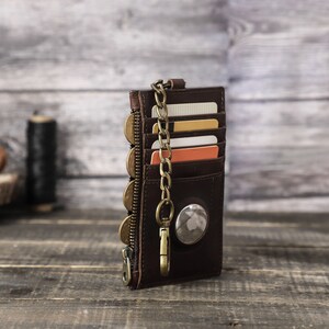 Puede incluir: Una cartera de cuero marr&oacute;n con cremallera y cadena. La cartera tiene m&uacute;ltiples ranuras para tarjetas y un bolsillo para monedas. La cartera est&aacute; sobre una superficie de madera.