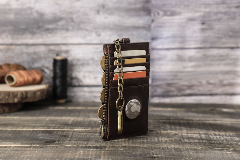 Puede incluir: Una cartera de cuero marr&oacute;n con cremallera y cadena. La cartera tiene m&uacute;ltiples ranuras para tarjetas y un bolsillo para monedas. La cartera est&aacute; sobre una superficie de madera.