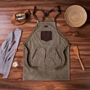 K&ouml;nnte beinhalten: Eine gr&uuml;ne Sch&uuml;rze aus Canvas mit braunen Lederriemen und Taschen. Die Sch&uuml;rze hat einen gro&szlig;en braunen Lederfleck auf der Vorderseite.