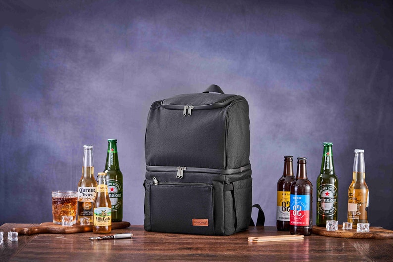 May include: A black insulated backpack cooler with two zippered compartments. The cooler is sitting on a wooden table with various bottles of beer and a glass of ice.