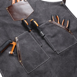 K&ouml;nnte beinhalten: Eine graue Canvas-Sch&uuml;rze mit braunen Lederverzierungen und Taschen. Die Sch&uuml;rze hat eine gro&szlig;e Tasche auf der Vorderseite mit zwei Schraubendrehern und einer Zange darin.