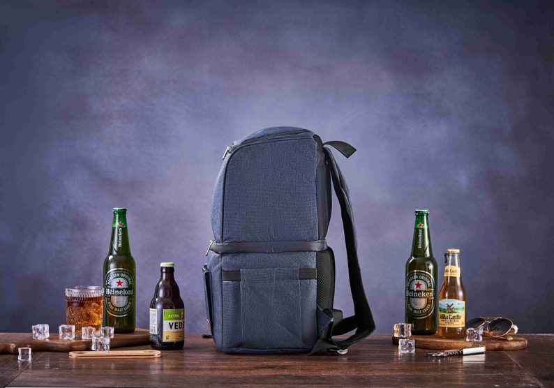May include: A blue insulated backpack cooler with adjustable straps. The cooler is on a wooden surface with four bottles of beer and a glass of beer.