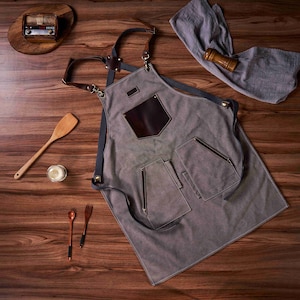 K&ouml;nnte beinhalten: Eine graue Leinensch&uuml;rze mit braunen Lederriemen und Taschen. Die Sch&uuml;rze hat einen gro&szlig;en braunen Lederfleck auf der Vorderseite mit einer kleineren Tasche dar&uuml;ber.