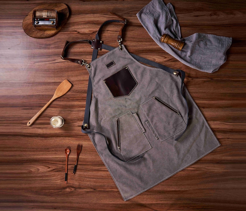 K&ouml;nnte beinhalten: Eine graue Leinensch&uuml;rze mit braunen Lederriemen und Taschen. Die Sch&uuml;rze hat einen gro&szlig;en braunen Lederfleck auf der Vorderseite mit einer kleineren Tasche dar&uuml;ber.
