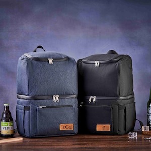 May include: Two insulated cooler backpacks, one blue and one black, with personalized leather patches. Both backpacks have two zippered compartments and a side pocket. The backpacks are sitting on a wooden surface with beer bottles and ice.