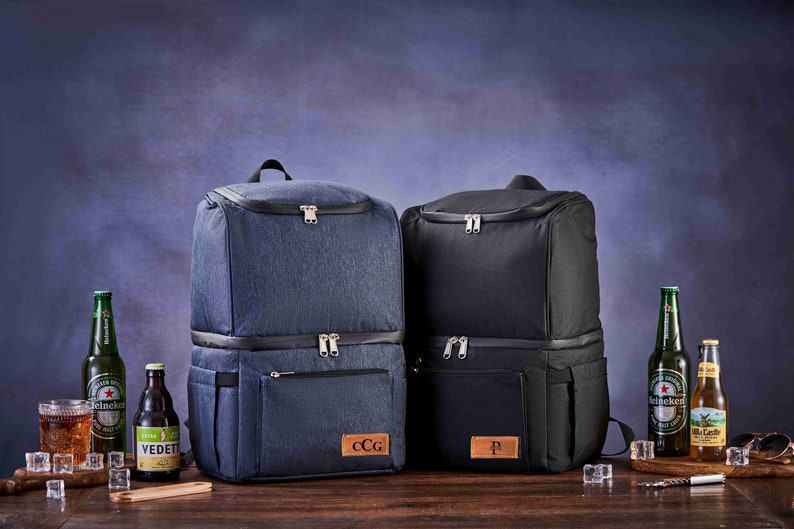 May include: Two insulated cooler backpacks, one blue and one black, with personalized leather patches. Both backpacks have two zippered compartments and a side pocket. The backpacks are sitting on a wooden surface with beer bottles and ice.