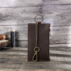 Puede incluir: Una cartera de cuero marr&oacute;n con cremallera y una cadena met&aacute;lica unida a un anillo. La cartera est&aacute; sobre una superficie de madera.