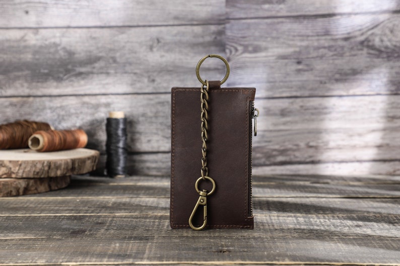 Puede incluir: Una cartera de cuero marr&oacute;n con cremallera y una cadena met&aacute;lica unida a un anillo. La cartera est&aacute; sobre una superficie de madera.