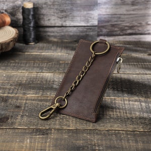 Puede incluir: Una cartera de cuero marr&oacute;n con cierre de cremallera y una cadena de lat&oacute;n unida a un llavero. La cartera est&aacute; sobre una superficie de madera.