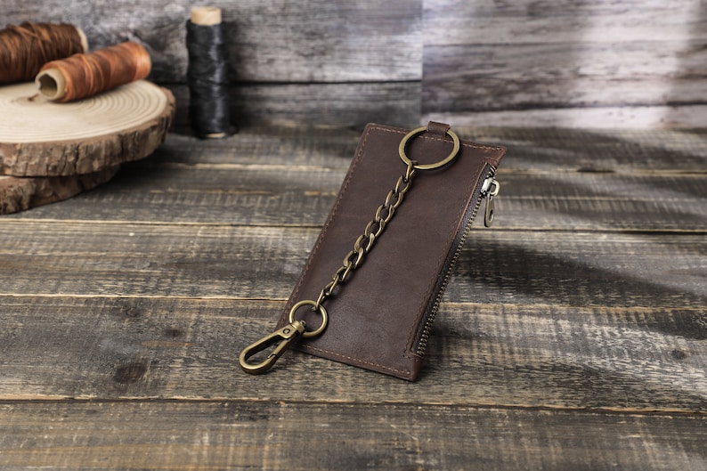 Puede incluir: Una cartera de cuero marr&oacute;n con cierre de cremallera y una cadena de lat&oacute;n unida a un llavero. La cartera est&aacute; sobre una superficie de madera.