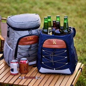 Peut inclure: Deux sacs à dos isothermes, un gris et un bleu marine, sont posés sur une table en bois. Le sac à dos gris porte les initiales "ERC" et le sac à dos bleu marine porte l'initiale "K". Les deux sont remplis de bouteilles de bière. Un refroidisseur de canettes et un ouvre-bouteille sont également sur la table.