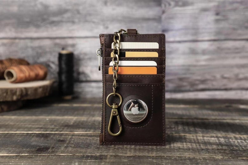 Puede incluir: Una cartera de cuero marr&oacute;n con cierre de cremallera y una cadena de metal. La cartera tiene m&uacute;ltiples ranuras para tarjetas y un peque&ntilde;o bolsillo para efectivo. La cartera tambi&eacute;n tiene un c&iacute;rculo de metal con un logotipo de Apple.