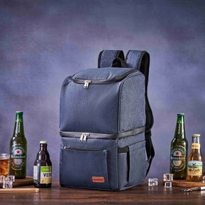 May include: A blue insulated backpack cooler with two zippered compartments and a front pocket. The backpack is on a wooden table with two bottles of beer, a bottle of brown liquid, and a glass of ice.