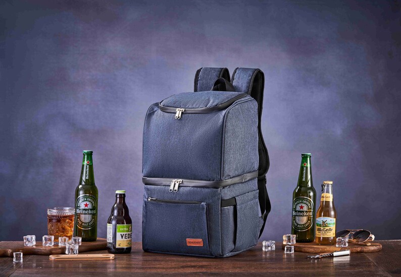 May include: A blue insulated backpack cooler with two zippered compartments and a front pocket. The backpack is on a wooden table with two bottles of beer, a bottle of brown liquid, and a glass of ice.