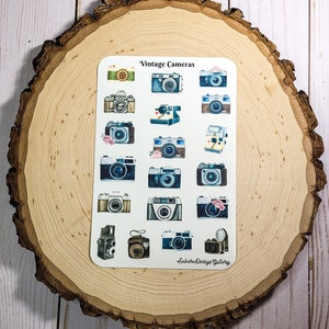 May include: A sheet of vintage camera stickers with a watercolor design. The stickers feature various styles of cameras, including a Polaroid, a 35mm film camera, and a vintage box camera. The stickers are on a white background and have a light blue and brown color scheme. The text "Vintage Cameras" is at the top of the sheet, and the text "Sakura Design Gallery" is at the bottom.
