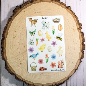 May include: A sheet of Easter themed stickers featuring a variety of images including a bunny, a lamb, chicks, a bee, a ladybug, a basket, flowers, and eggs. The stickers are on a white background and have a watercolor look. The text "Easter" is at the top of the sheet and the text "Sakura Design Gallery" is at the bottom.