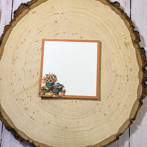 May include: A square card with an orange border and a fall-themed illustration. The illustration features sunflowers, pumpkins, and gourds in and around wooden barrels. The card is set on a large, round, wooden slice.